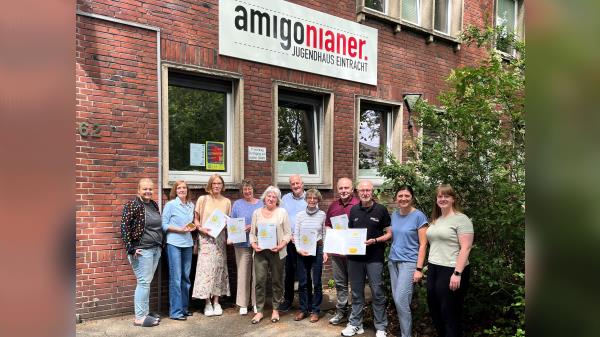 Foto von einer Gruppe Menschen vor einem Gebäude mit dem Schriftzug Amigonianer. Sie halten Mappen mit der Ehrenamtskarte NRW in den Händen