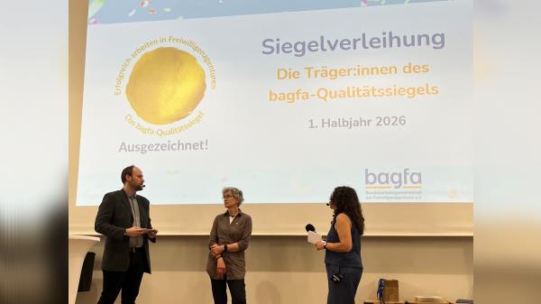 Foto der Siegelverleihung mit drei Personen, im Hintergrund wird mit einem Beamer der Schriftzug Siegelverleihung an die Wand geworfen.